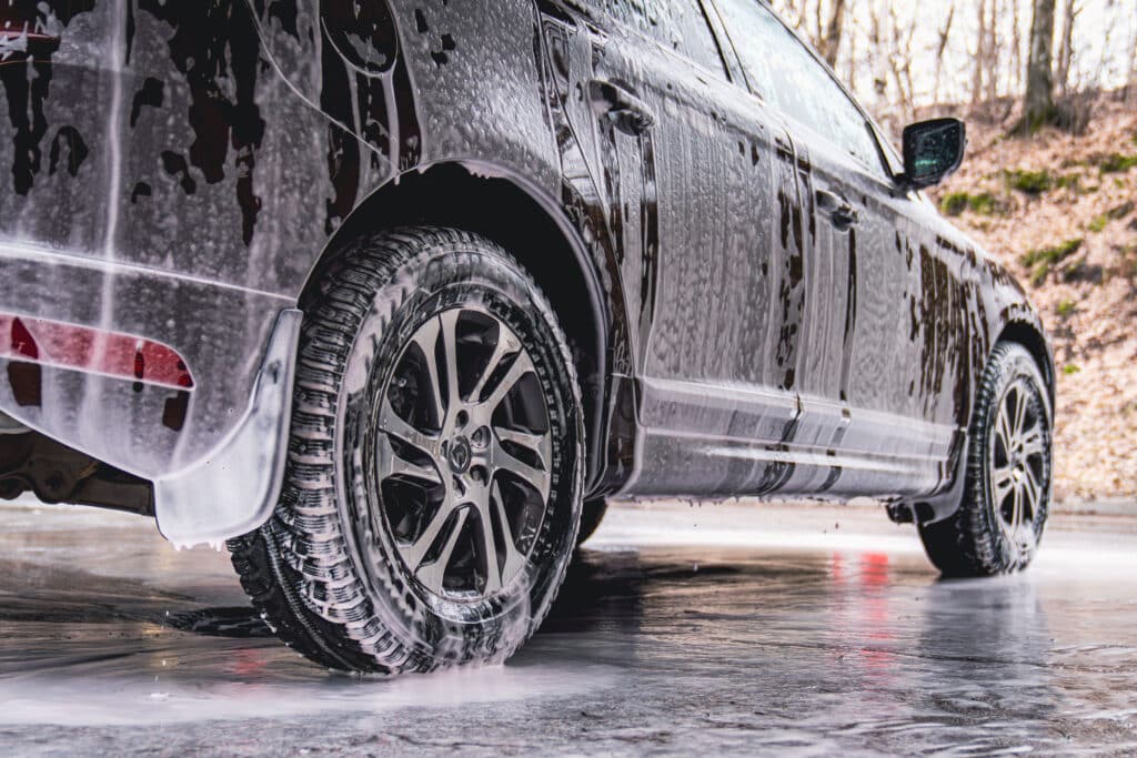 Vehicle covered in white soapy foam during regular carwash outdoors, auto getting professional wash with soap at washing center, cleaning car exterior surface at self-service station