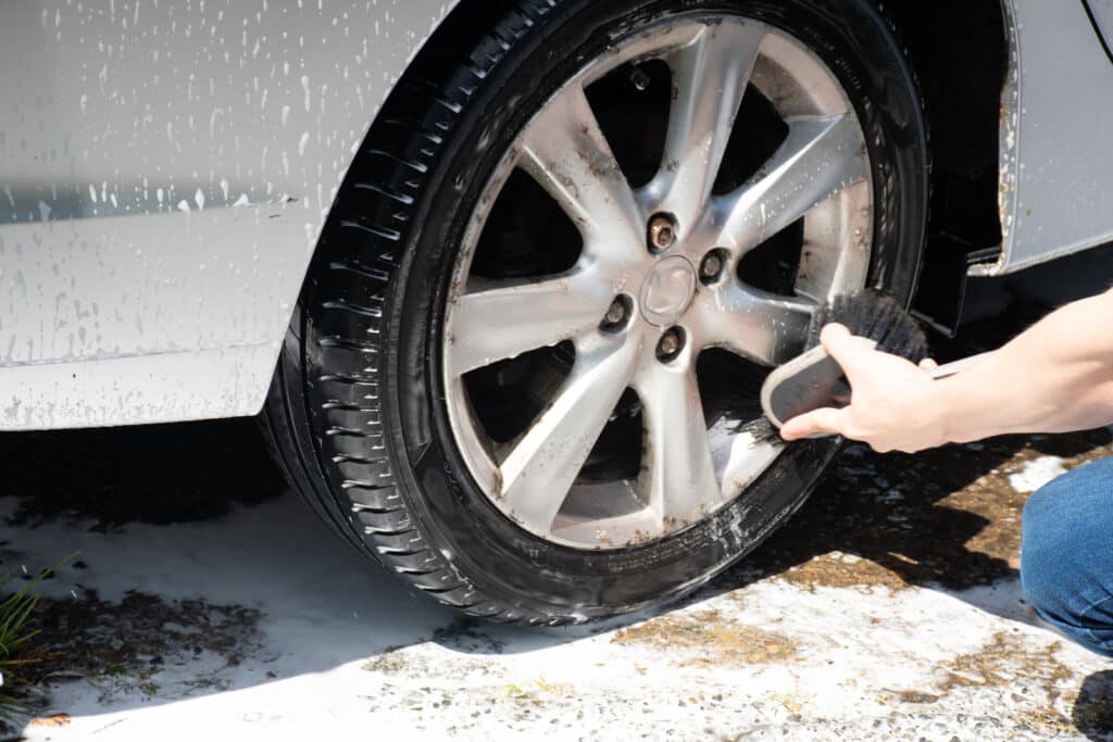 Nettoyage jante et pneu de voiture sale au savon Bras d'une personne lavant une jante de voiture sale avec une brosse noire, mousse et eau savonneuse sur le pneu et la carrosserie grise.
