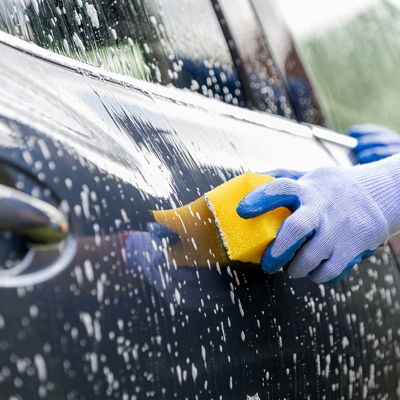Lavage voiture : main gantée avec éponge jaune Main gantée de bleu lavant la carrosserie noire d'une voiture avec une éponge jaune et de la mousse savonneuse.