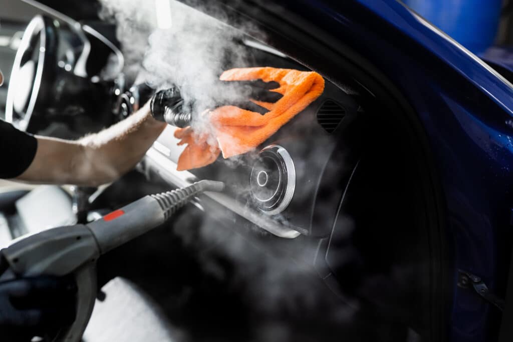 Nettoyage Vapeur Intérieur Auto : Détachage Détaillé Un technicien ganté nettoie la console de voiture bleue avec de la vapeur et un chiffon microfibre orange vif, détachage intérieur.