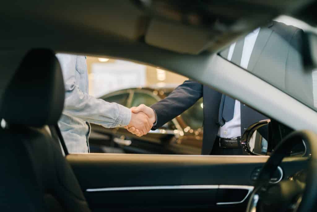 Poignée de main achat voiture : conclusion affaire Poignée de main entre un homme en chemise en jean et un homme d'affaires en costume, vus de l'intérieur d'une voiture dans un concessionnaire.